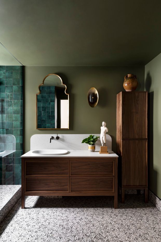 A richly styled bathroom featuring a dark walnut floating vanity with integrated white basin and rounded terrazzo-like benchtop, deep olive-green walls, teal square wall tiles in the shower area, and a decorative scalloped mirror with brass-toned edging. A matching tall timber storage cabinet, sculptural wall light, pottery, plant, and figurine add a refined, retro-inspired character. The floor is finished in terrazzo with dark and light aggregate.