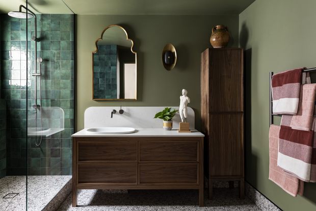 A moody, refined bathroom with a walk-in glass shower clad in deep green square tiles, a freestanding-style double-drawer timber vanity with white basin, and a tall dark timber storage cabinet. An ornate arched mirror, sculptural wall light, potted plant, and burgundy-and-white towels add decorative character. The room features terrazzo-style flooring and dark olive-green painted walls.