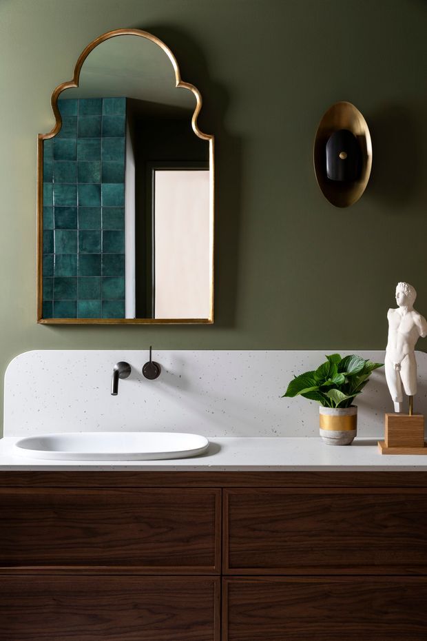 A richly styled bathroom vanity with dark walnut cabinetry, a white speckled stone benchtop and integrated splashback, a white vessel basin, dark brushed-metal tapware, and deep olive-green walls. An ornate scalloped bronze-framed mirror reflects teal tiled wall detailing and a doorway. A sculptural wall light, potted plant, and small classical statue add decorative character.