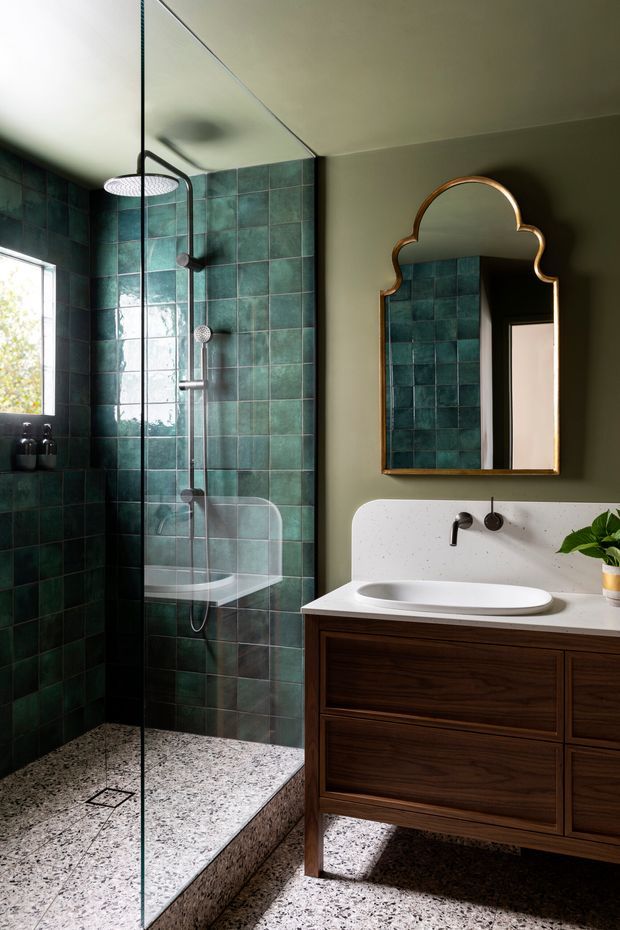 A sophisticated bathroom with a walk-in glass shower lined in deep green square tiles, a terrazzo floor and shower base, and a dark timber floating vanity with a white integrated basin. A sculptural brass-framed mirror, black tapware, and olive-green walls add a rich, moody character.
