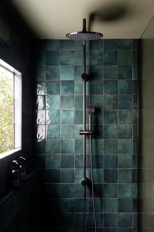 A moody, compact bathroom shower area featuring dark teal-green glazed wall tiles arranged in a regular grid, black tiled side walls, and a matte dark metal rain shower with handheld attachment. Natural light enters through a small window on the left, creating reflections on the glossy tiles.