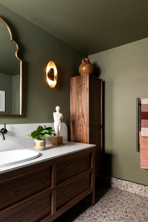 A moody, refined bathroom featuring a dark green painted wall and ceiling, a large sculptural framed mirror, a warm wall sconce, a white countertop with an integrated basin, and dark timber vanity cabinetry. A tall matching timber storage cabinet, terrazzo-style flooring, decorative pottery, and a heated towel rail with blush and cream towels complete the space.