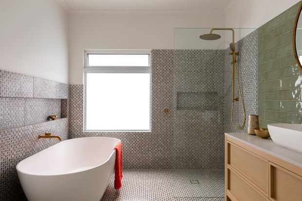 Bright contemporary bathroom with a freestanding white bathtub, walk-in shower, wall-mounted brass fixtures, white basin, and timber vanity cabinetry. Small patterned mosaic tiles cover the floor and much of the walls, contrasted with glossy muted green rectangular tiles in the shower area. A large window provides natural light.