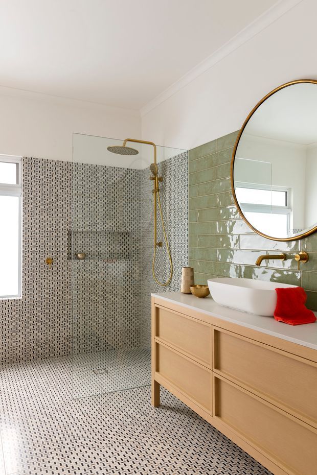 Bright contemporary bathroom featuring a walk-in shower with a rainfall showerhead and handheld fitting in brushed brass, small patterned mosaic wall and floor tiles, a glossy sage-green tiled vanity wall, and a light timber floating vanity with a white vessel basin. A large round brass-framed mirror, glass shower screen, and a red hand towel add detail.
