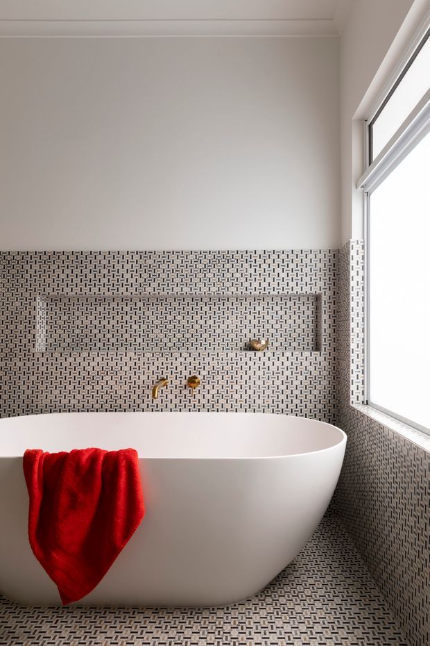 A bright, refined bathroom featuring a freestanding white bathtub, patterned mosaic tile covering the floor and lower walls, a recessed tiled shelf, brushed gold wall-mounted tapware, and a large white-framed window. A vivid red towel adds contrast.