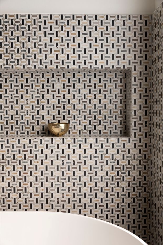 Close-up view of a sophisticated bathroom feature wall clad in intricate geometric mosaic tiles. The patterned tiles cover the walls and recessed shelving, with a small metallic bowl on the shelf and the curved edge of a white freestanding or built-in bathtub visible in the foreground. The palette is predominantly white, cream, grey, taupe, and black with subtle metallic accents.