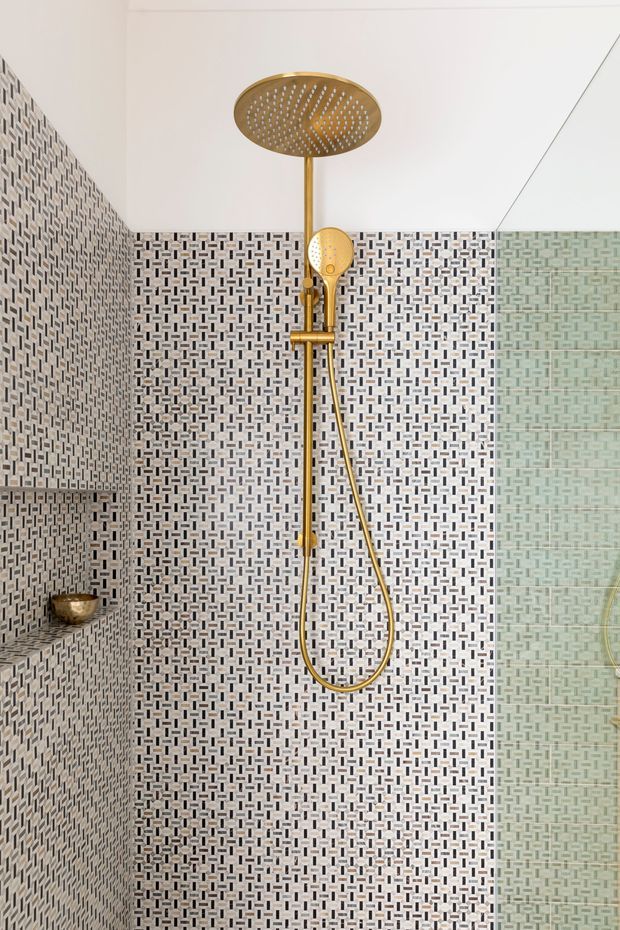 Close-up of a contemporary shower enclosure with a large round gold rain showerhead, matching handheld shower, and exposed riser rail. The walls are covered in a small repeating geometric mosaic tile pattern in white, grey, and charcoal, with a recessed tiled shelf and gold fixture visible on the left. A frameless glass screen appears on the right.