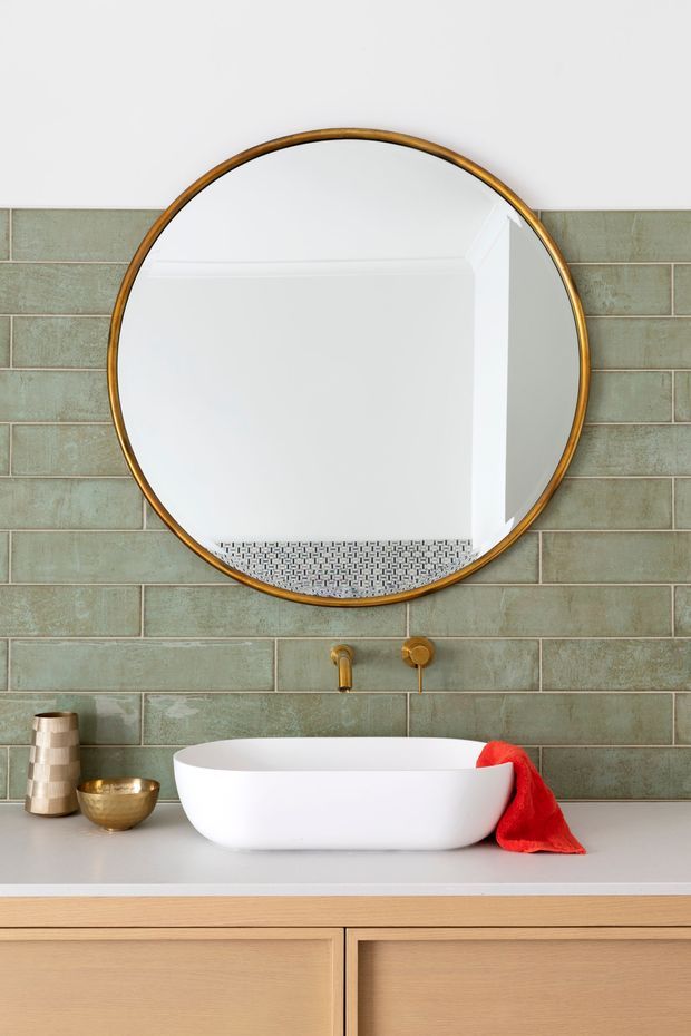 A styled bathroom vanity featuring a white freestanding vessel basin on a light timber floating cabinet, aged green rectangular wall tiles, and brushed brass wall-mounted tapware. A large round mirror with a brass frame dominates the wall, with small decorative vessels and a red towel adding accents.