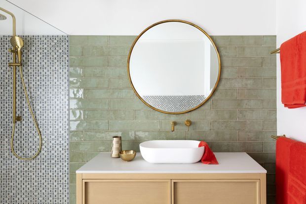 A bright contemporary bathroom vanity with a white vessel basin on a light timber double-drawer vanity. The wall is clad in muted sage-green rectangular tiles, with a contrasting small geometric mosaic tile shower wall. Brass shower fittings, tapware and round mirror frame add warm metallic accents, while vivid red towels provide colour contrast.