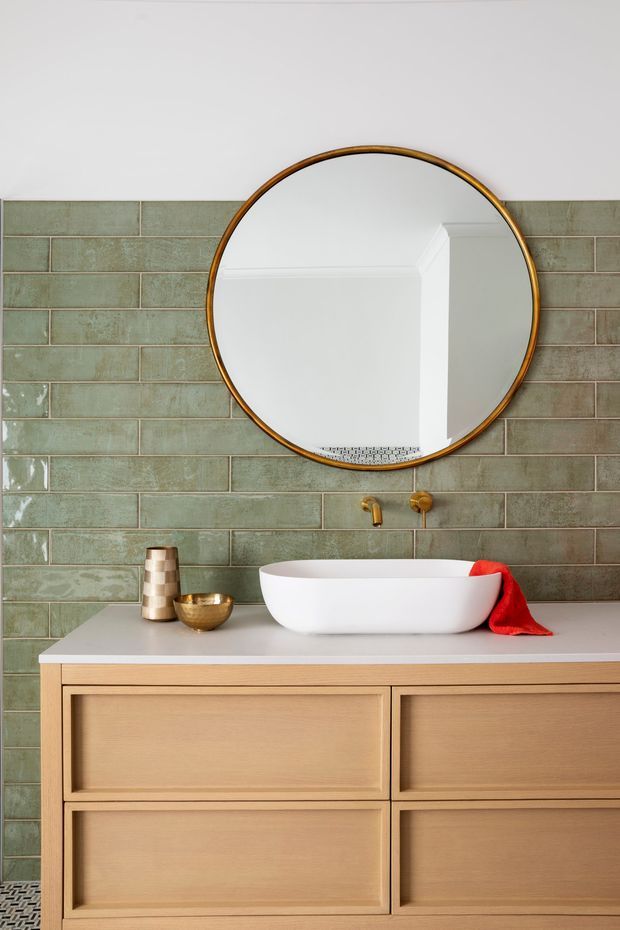 A refined bathroom vanity vignette featuring a large round brass-framed mirror, pale sage-green rectangular glazed wall tiles with contrasting grout, a white vessel basin on a light timber vanity, and brushed brass wall-mounted tapware. Small decorative accessories and a red cloth add colour. A patterned tile floor is partially visible at the bottom.