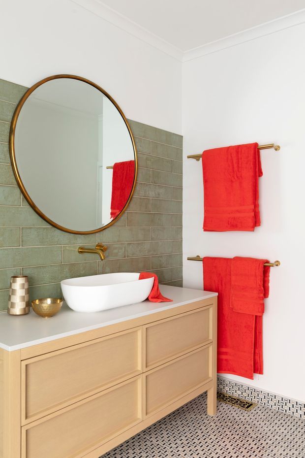 A bright, refined bathroom featuring a floating-style light timber vanity with framed drawer fronts, a white vessel basin, brushed brass tapware and towel rails, a large round brass-framed mirror, muted green rectangular wall tiles, and a patterned small-format tiled floor. Bold red towels provide a vivid accent against the white walls.