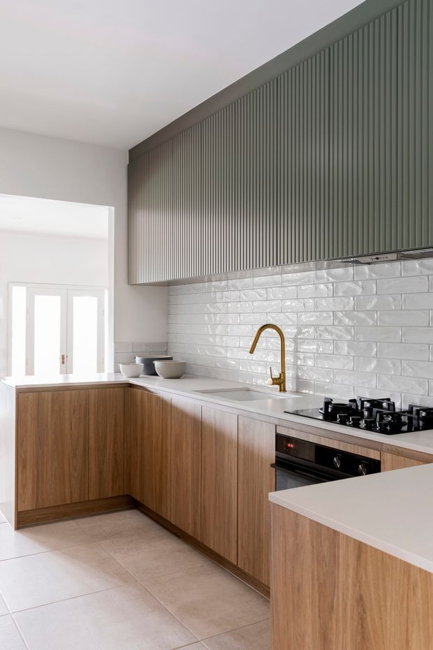 Bright contemporary kitchen with an L-shaped timber-look cabinetry layout and peninsula, white engineered stone benchtops, glossy white subway-tile splashback, vertically fluted muted green-grey upper cabinetry, and a brass gooseneck tap. A freestanding cooker and oven sit beneath the rangehood, with a glazed door bringing in natural light.