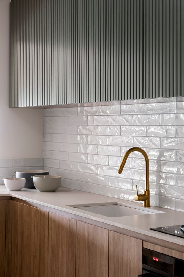 Contemporary compact kitchen with vertically ribbed grey upper cabinetry, glossy white rectangular tiled splashback, pale benchtop with integrated sink, warm timber lower cabinetry, and a curved brass-gold mixer tap. Neutral, refined finishes create a clean modern look.