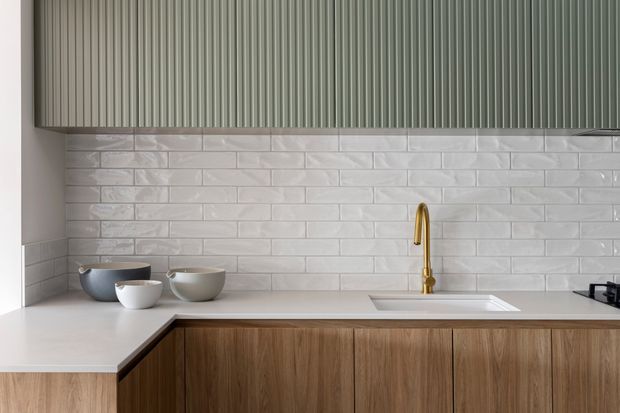 Contemporary kitchen detail featuring warm vertical-grain timber cabinetry, a pale benchtop, glossy white subway-tile splashback, and a brushed gold gooseneck tap. Ribbed vertical cabinetry or panelling appears above the splashback, with ceramic bowls on the counter and a partially visible cooktop at right.