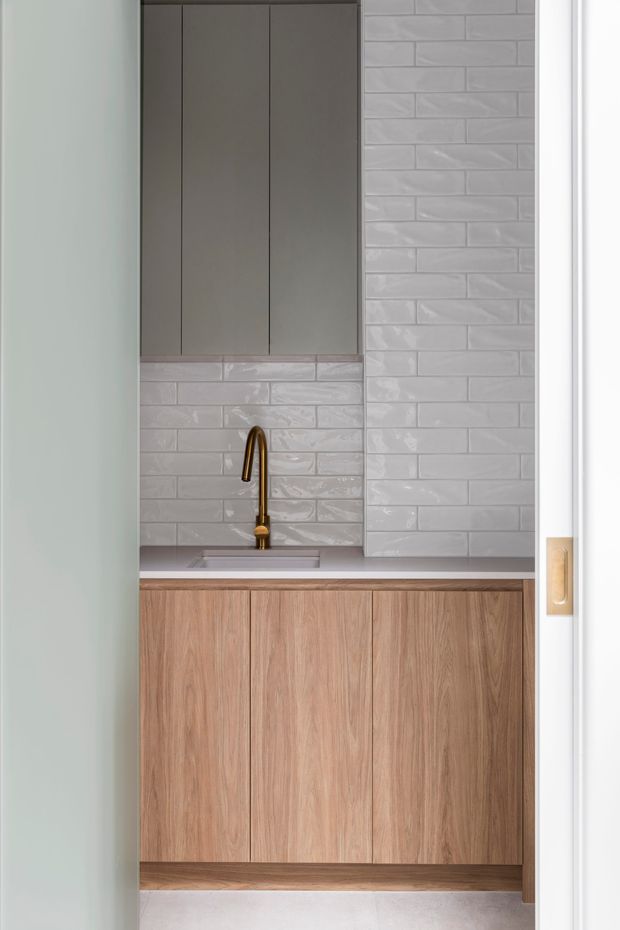 Compact, modern utility or kitchenette space viewed through a doorway, featuring warm wood-grain base cabinetry, dark taupe wall cabinetry, a pale grey engineered-stone benchtop, glossy light-grey subway tile splashback, and a brass gooseneck tap over a small integrated sink.