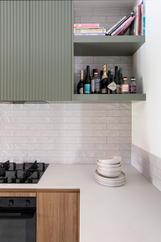 A close-up view of a contemporary kitchen corner featuring pale grey subway-tile splashback walls, a light neutral benchtop, warm timber lower cabinetry, and muted green vertically fluted upper cabinetry. An integrated open shelf displays books and bottles, while a black gas cooktop and oven occupy the lower left. Stacked white bowls and plates sit on the benchtop.