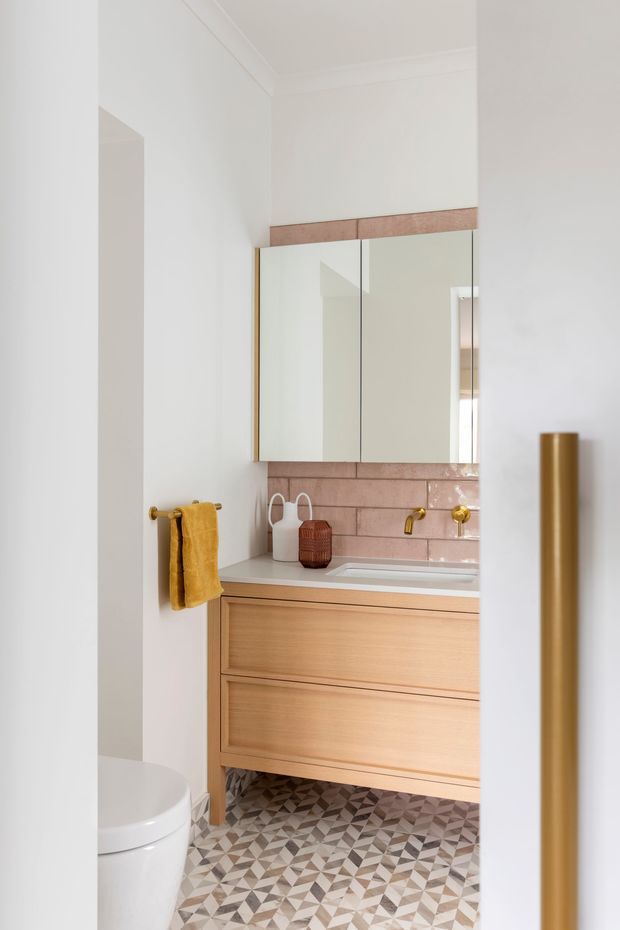 A compact, refined bathroom viewed through an open doorway. The room features a wall-mounted timber vanity with integrated basin, mirrored wall cabinet, pink-toned tiled splashback, gold tapware and towel rail, and a patterned geometric tiled floor. A toilet is partially visible in the foreground, while white walls and ceiling cornices create a bright, polished setting.