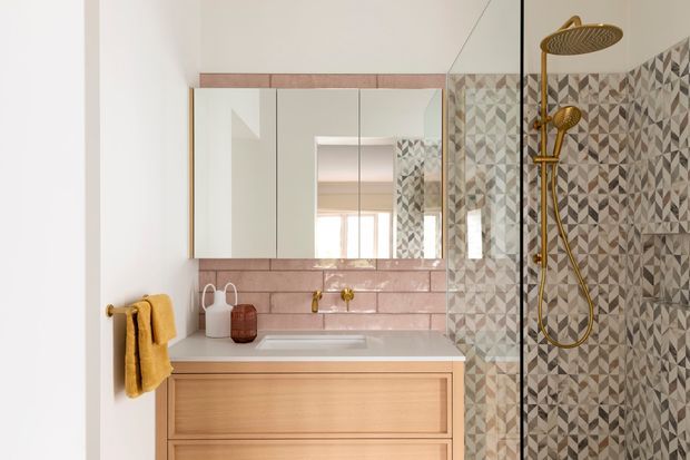 A compact, brightly lit bathroom featuring a timber vanity with integrated basin, mirrored wall cabinets, blush-pink rectangular wall tiles, and a patterned geometric tiled shower enclosure. Gold-toned tapware, shower fittings, and towel rail add a warm accent.