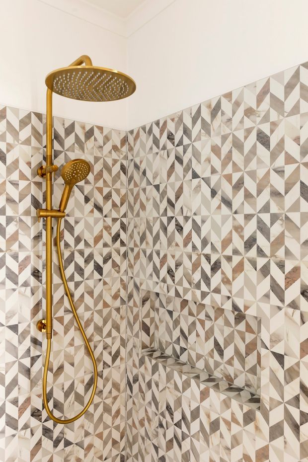 Close-up view of a contemporary shower enclosure featuring a large rainfall showerhead and handheld shower in a warm brass finish. The walls are clad in a striking geometric patterned tile composed of cream, beige, grey, taupe, and brown triangular forms, with a built-in tiled niche visible in the corner.