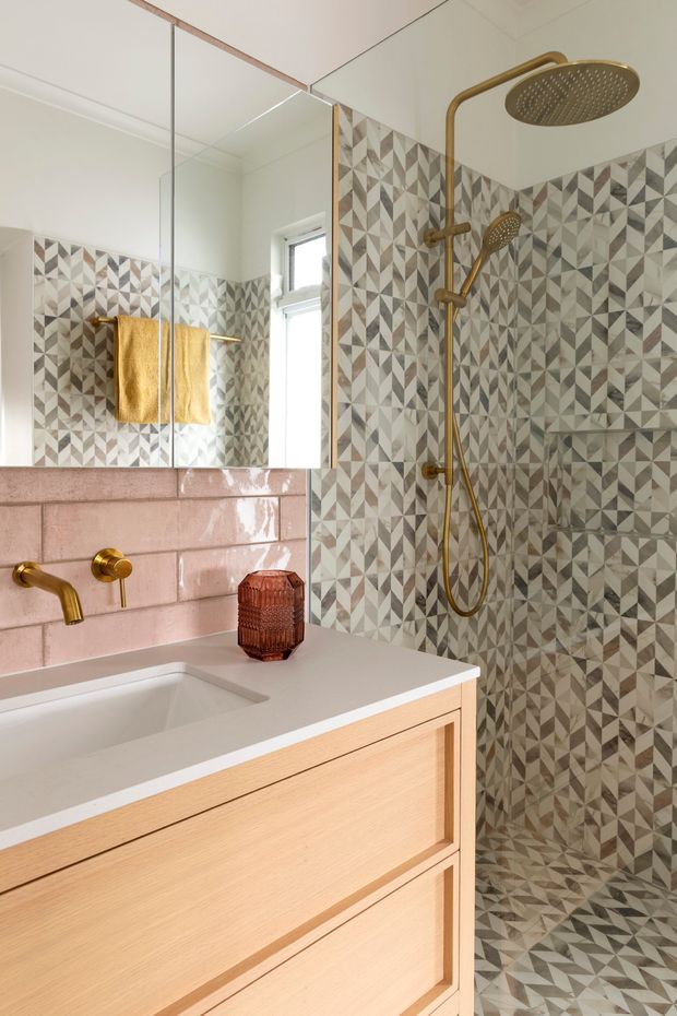 A compact, contemporary bathroom featuring a walk-in shower clad in geometric patterned tiles, a glossy blush-pink tiled wall, a pale timber vanity with a white benchtop and integrated basin, and brass/gold tapware and shower fittings. A large mirror and warm yellow towels add brightness and contrast.