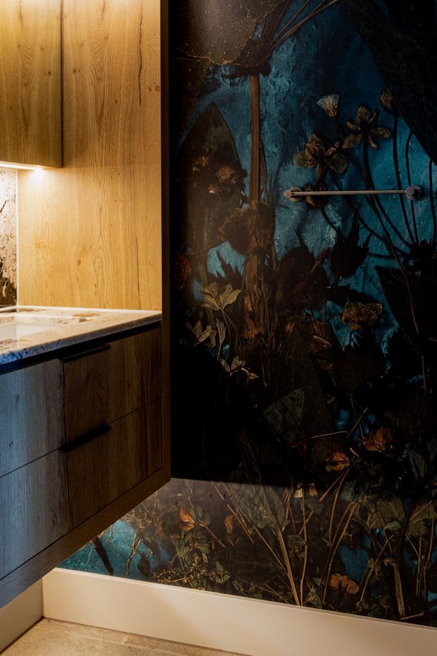 A moody bathroom detail featuring warm timber-grain wall cabinetry with integrated lighting, a pale stone benchtop, and dark blue botanical patterned wall surfaces extending around a bathtub. A slim horizontal metal towel rail is mounted on the feature wall, and the floor has a neutral stone or tile finish.