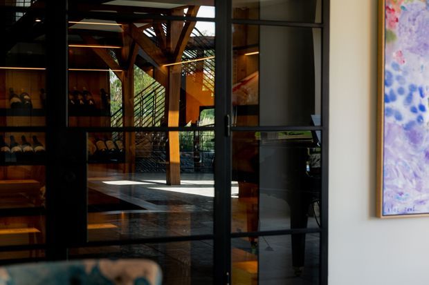 Interior view through black-framed glazed doors into a sophisticated timber-structured space. The room features exposed timber beams and columns, dark reflective flooring, extensive glazing, warm linear lighting, shelving or cabinetry, and a colourful artwork on the right wall. The image suggests a refined entry or transitional area opening toward a larger interior or courtyard-like space.