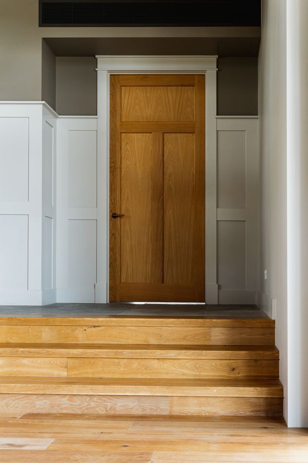 Interior view of a traditional-style stair landing with broad warm timber stairs leading up to a paneled timber door. The door is framed by white decorative trim and flanked by white built-in panelled cabinetry or wainscoting. Dark grey painted walls and a black linear ventilation grille create contrast, while natural timber flooring and steps add warmth.