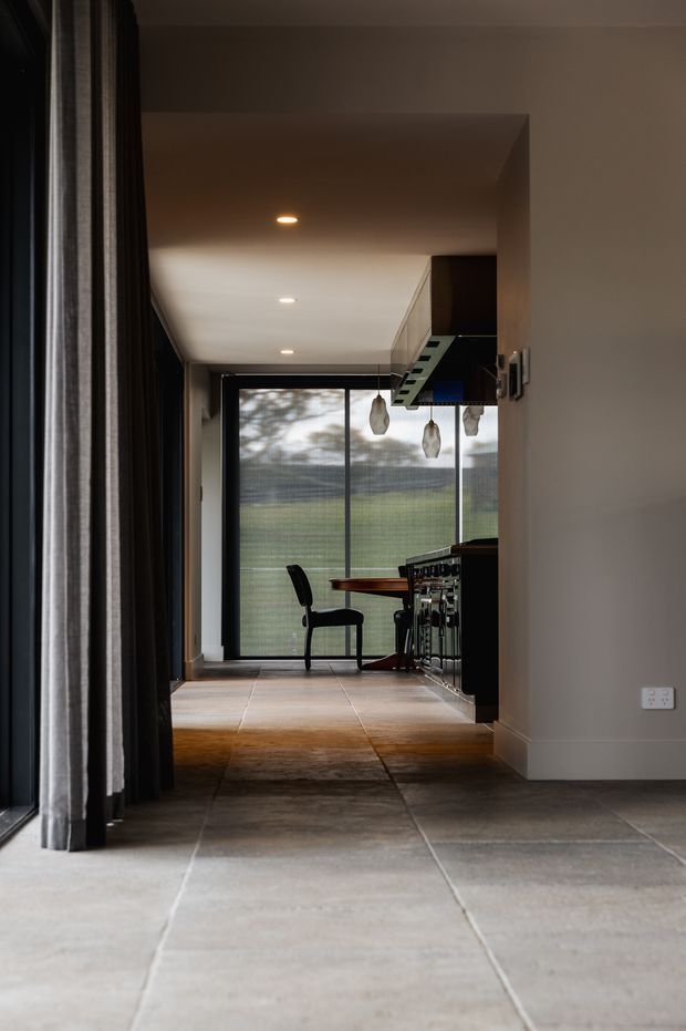 A dark, refined interior view through a hallway toward a compact kitchen or bar area. Large grey-format floor tiles lead to warm timber flooring, with dark cabinetry, a black-framed glazed door or window, pendant lights, and a single chair. Floor-to-ceiling curtains line the left side, while the space beyond opens to a green outdoor view.