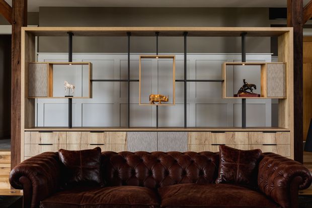 Interior view of a refined living room dominated by a deep brown tufted leather Chesterfield sofa in front of a large custom-built timber and black metal display joinery wall. The joinery includes open glass-fronted niches, integrated lighting, decorative objects, and lower wood-grain cabinetry. Vertical timber structural elements and a neutral paneled wall create a warm, tailored, slightly industrial character.