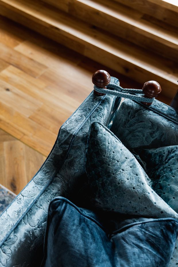 Close-up detail of a richly upholstered dark teal-blue textile draped over a wooden stair or railing, secured with a dark rope and polished wooden finials. Warm timber flooring and timber stair elements form the background, creating a tactile, moody interior composition.