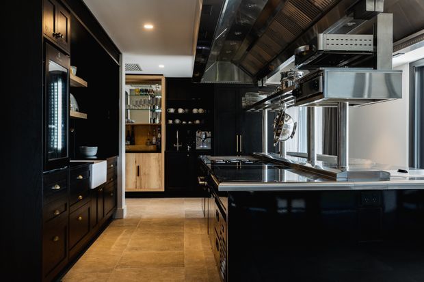 A dramatic, high-end galley kitchen with glossy black cabinetry, warm timber shelving and cabinetry, large stainless-steel cooking and extraction equipment, and a broad central island. Tan stone-look flooring, reflective metal surfaces, integrated lighting, and extensive glazing create a refined professional-kitchen atmosphere.