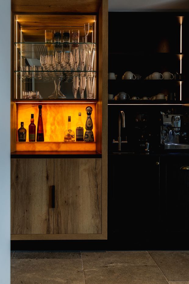 A dark, refined home bar and beverage station featuring a timber-framed illuminated display cabinet with glass shelving, crystal stemware, and liquor bottles. Adjacent black cabinetry contains open shelving with cups, a sink with brass-toned tapware, and an integrated espresso machine. The lower cabinetry is timber veneer with long black handles, while the floor is finished in large rustic stone-look tiles. Warm amber lighting creates a luxurious, intimate atmosphere.