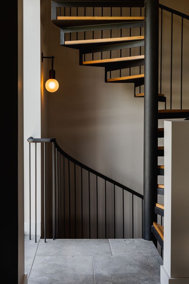 A dimly lit interior stairway featuring a sculptural spiral or switchback stair with dark metal balustrades and central post, warm timber stair treads, a grey stone or concrete landing floor, and a small glowing pendant light. The composition emphasizes the dramatic stair geometry and layered vertical circulation.