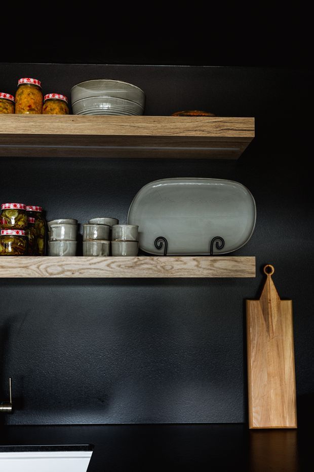 Close-up of a dark, moody kitchen wall with two open timber shelves displaying stacked ceramic dishes, glass jars and preserves. A large serving platter rests on the lower shelf, while a timber cutting board hangs beside it. The lower edge reveals part of a black benchtop and a white ceramic sink or appliance.