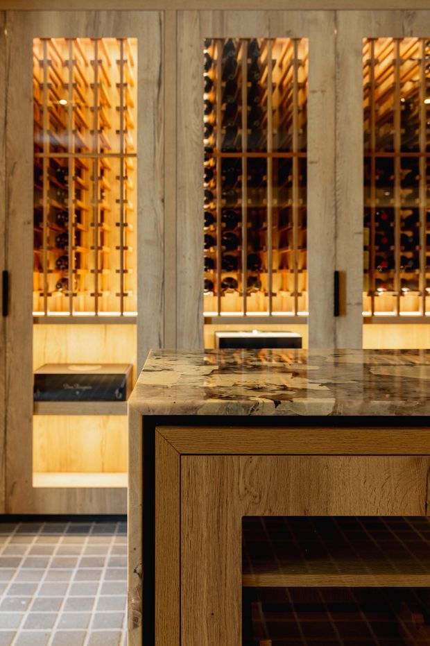 A warm, refined wine room with floor-to-ceiling timber wine racks behind a vertical timber screen. The foreground features a substantial natural stone island or counter with integrated timber cabinetry, while small dark floor tiles and concealed amber lighting reinforce the intimate cellar atmosphere.