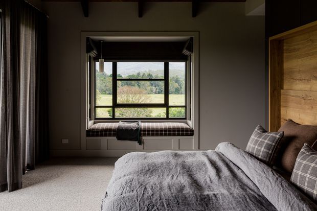 A moody, refined bedroom with a large framed window and built-in window seat overlooking a green landscape. The room features exposed dark ceiling beams, neutral painted walls, full-height dark curtains, plush carpet, and a bed with grey textured bedding and plaid cushions. A warm timber panel forms the headboard wall, adding natural contrast.