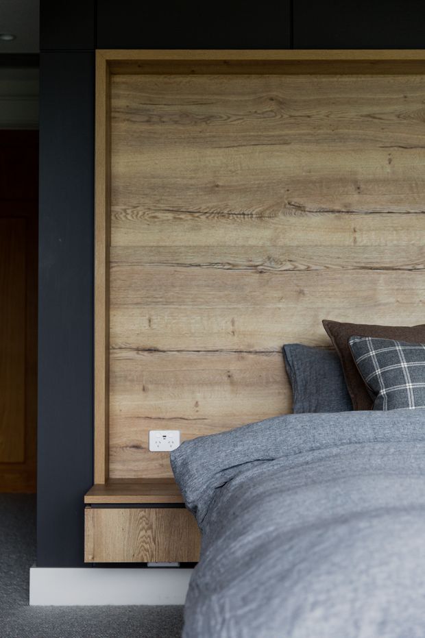 A close-up view of a contemporary bedroom featuring a bed with layered grey bedding and plaid cushions, a large framed natural timber feature wall behind the bed, and an integrated timber bedside drawer. Dark charcoal wall panels and trim create contrast, with carpeted flooring and a white power outlet visible beside the bed.