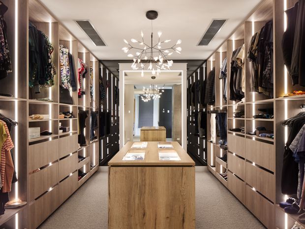 A spacious walk-in wardrobe with symmetrical floor-to-ceiling timber-look cabinetry, open hanging rails, drawers, integrated vertical lighting, and a central timber island. A large mirror at the far end reflects the room, while a sculptural branch-like pendant light hangs from the ceiling. The room has a soft neutral palette and carpeted floor.