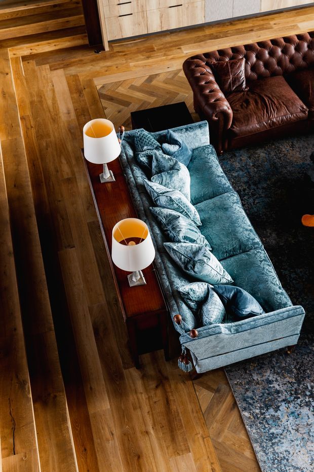 Overhead view of a richly styled living room featuring a blue patterned chaise-style sofa with layered cushions, a deep brown tufted leather sofa, two white table lamps on a dark wood console, and warm timber flooring with a geometric parquet inset. A patterned area rug anchors part of the seating area.