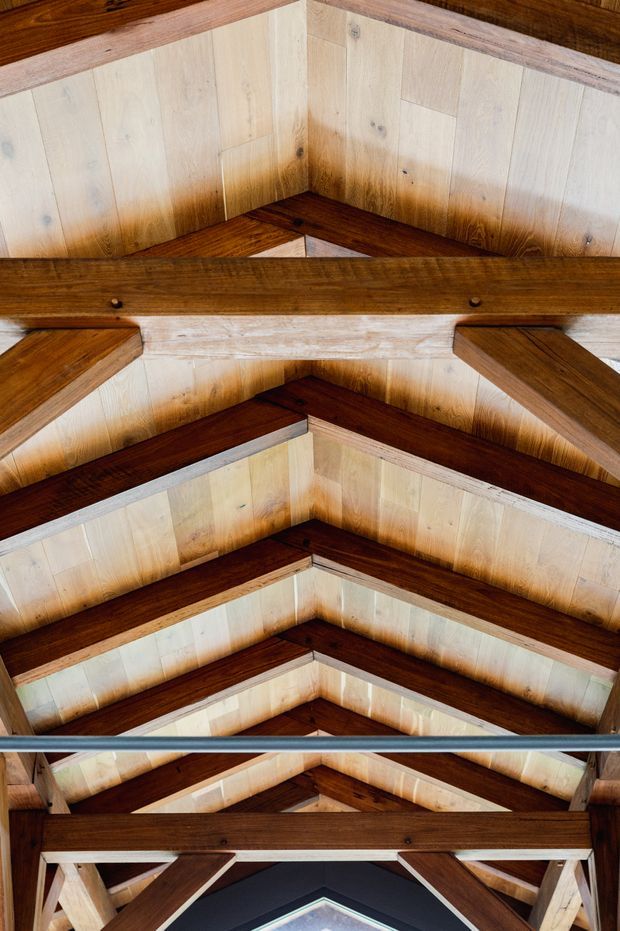 Upward-looking view of a timber roof structure, showing repeated dark exposed rafters and beams against pale timber roof lining, with a central triangular gable-like geometry and a small glimpse of glazing or sky at the bottom. The image focuses on exposed structural carpentry rather than a furnished room.