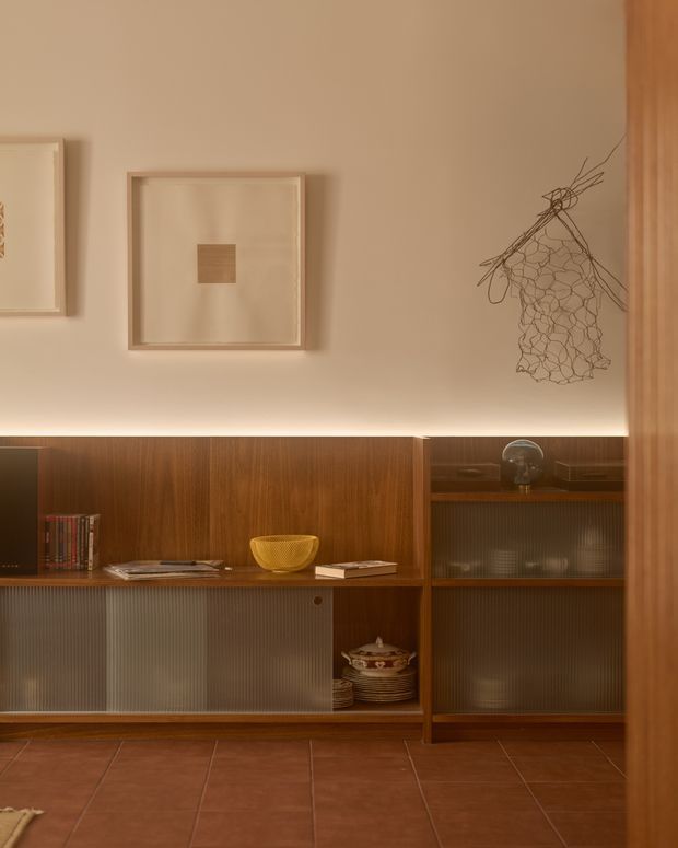 Warmly lit interior vignette featuring a full-height dark timber built-in storage and display unit with fluted translucent sliding panels, open shelving, books and decorative objects. The upper wall is painted in a soft beige tone with framed artwork and a delicate sculptural wall piece. Terracotta-toned tiled flooring and timber joinery create a refined, mid-century-inspired atmosphere.