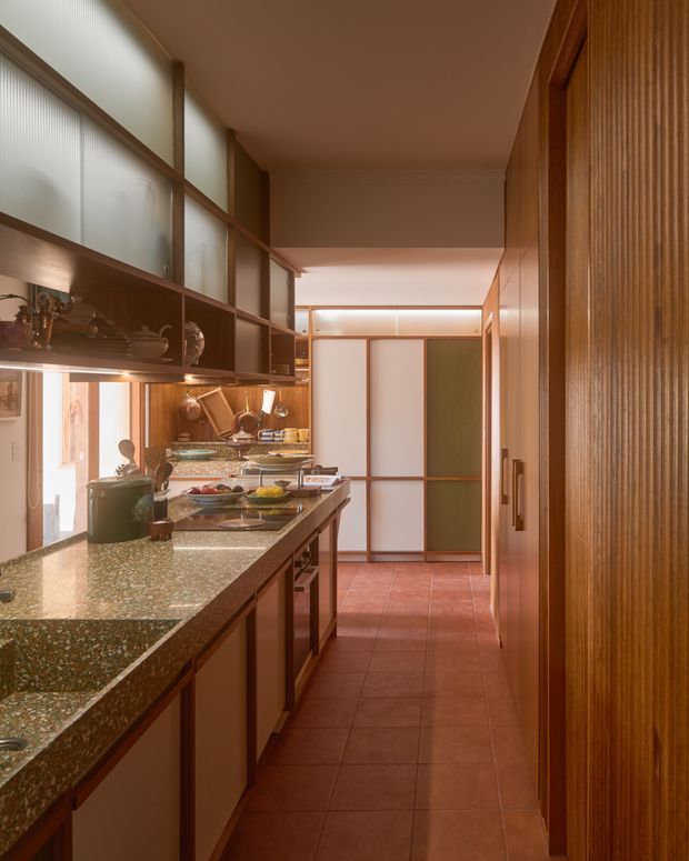 A narrow galley kitchen with warm wood cabinetry and wall panelling, a speckled stone benchtop, integrated open shelving, and translucent glass-panelled upper sections. Terracotta-toned tiled flooring, copper-toned accents, and warm concealed lighting create a richly layered, mid-century-inspired interior. The long circulation axis extends toward glazed and timber-framed doors at the rear.