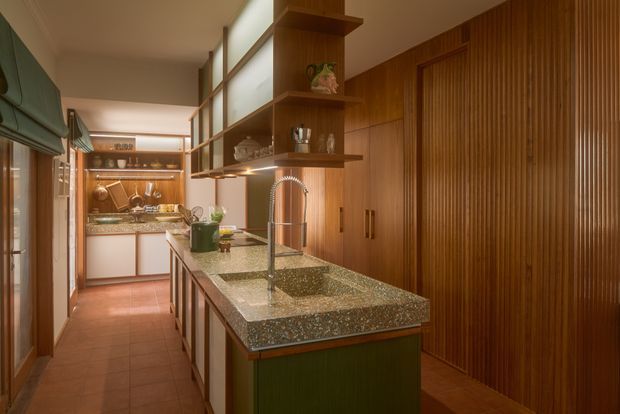 A warm, narrow galley-style kitchen with a central island featuring a speckled stone benchtop, double undermount sinks and a tall gooseneck tap. Rich timber wall panelling and open shelving create a strong built-in feature, while green cabinetry, terracotta-toned floor tiles, and bronze-toned fittings add colour and warmth. A secondary kitchen workspace is visible at the rear, with glazing and blinds along the left side.