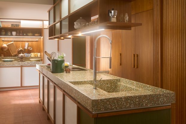 Warmly lit kitchen centred on a large island with an integrated double-bowl sink and tall chrome gooseneck tap. The island has a heavily patterned stone benchtop and white panelled cabinetry with timber edging. The surrounding kitchen features extensive dark timber cabinetry, open display shelving, vertical fluted timber detailing, under-cabinet lighting, and a tiled floor. The composition has a distinctly retro-inspired, refined kitchen fit-out.