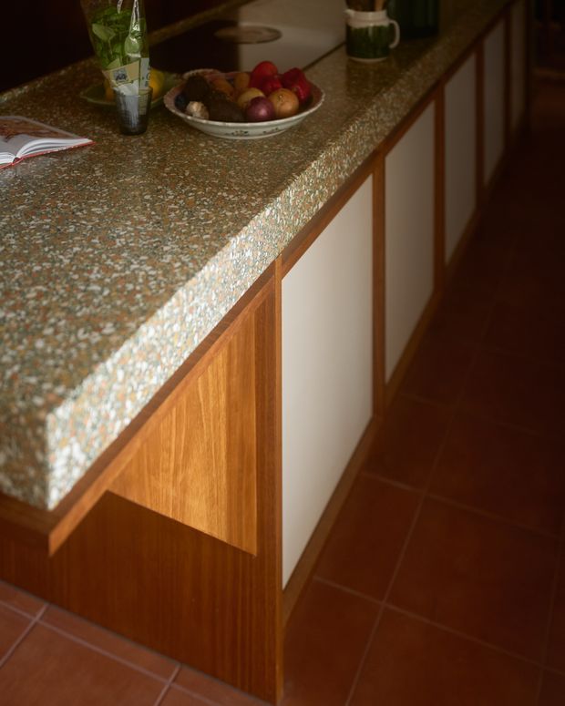 Close-up view of a retro-style kitchen island or peninsula with a thick terrazzo countertop, warm timber cabinetry, and contrasting cream inset cabinet panels. Terracotta floor tiles and a bowl of fruit reinforce the warm, vintage character.