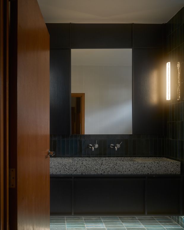 A dark, moody contemporary bathroom viewed through a partially open timber door. The room features glossy dark tiled walls, a large frameless rectangular mirror, wall-mounted basin fixtures, a substantial speckled terrazzo benchtop, matte dark built-in cabinetry, blue-green floor tiles, and a slim vertical wall light.