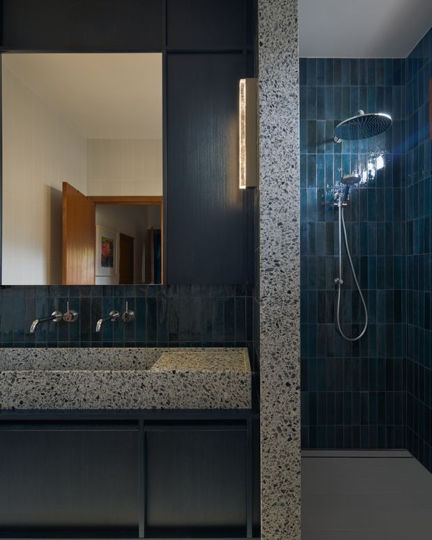 A dark, contemporary bathroom vanity and shower area. The room features deep blue vertically stacked wall tiles, a terrazzo vanity benchtop and matching structural divider, dark charcoal cabinetry, integrated warm strip lighting, and a large frameless mirror. A rain shower and handheld shower are visible in the tiled enclosure.