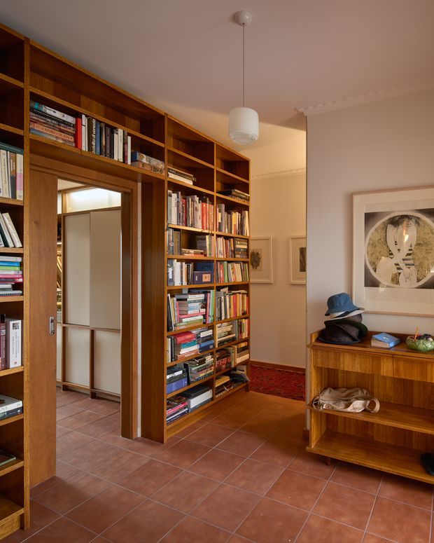 Warm interior view of a residential hallway or entry area lined with extensive built-in timber bookcases and open shelving. A timber-framed doorway leads to another tiled space, while a low timber console with shelving sits against the wall beneath framed artwork. Terracotta-toned square floor tiles, cream walls, decorative ceiling cornices, and a white pendant light create a richly furnished, book-filled atmosphere.