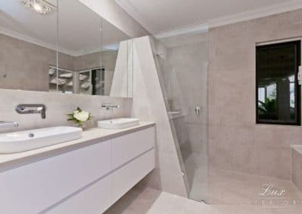 Bright contemporary bathroom with a long floating white vanity, twin basins, wall-mounted chrome tapware, large mirrored wall, and pale stone-look tiled walls and floor. A frameless glass shower sits behind a sculptural angled partition, with a black-framed window and partial bathtub visible on the right.
