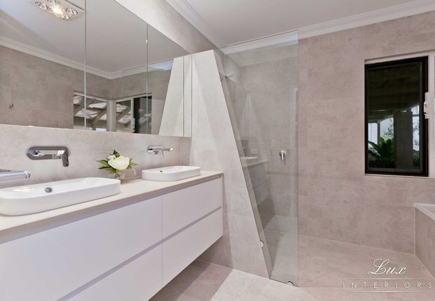 Bright contemporary bathroom with double white vessel basins on a floating vanity, wall-mounted chrome tapware, large mirrored cabinetry, pale stone-look tiled walls and floor, and a frameless glass shower enclosure. A freestanding or partially visible bathtub appears at the right edge, with a black-framed window providing natural light.